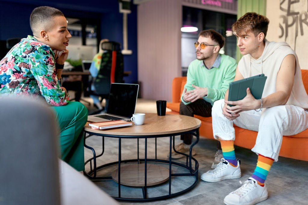 Trois jeunes adultes échangent de façon détendue dans un espace de travail; l'un porte des chaussettes aux couleurs de l'arc-en-ciel.